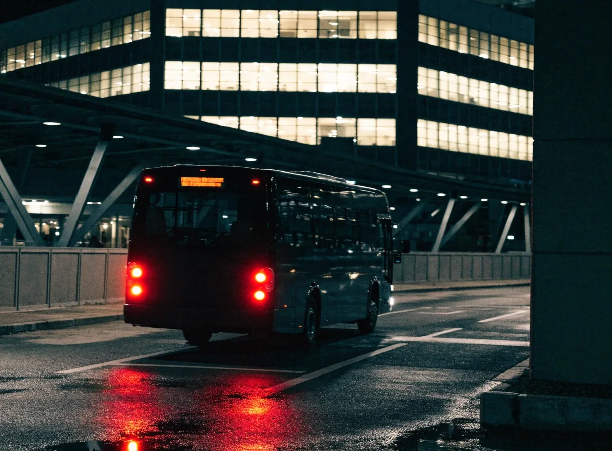 A courtesy bus pulling away from a kerbside stop on a short parking driveway at night, tail lights on wet tarmac